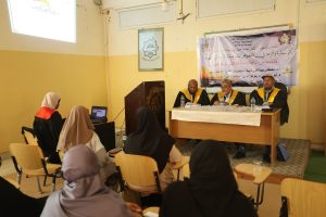 Panel discussion in a classroom: three men in robes with yellow sashes sit at a table at the front, Arabic banner behind them, audience of hijab-wearing women in chairs.