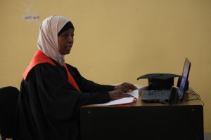Portrait of a woman in a black graduation gown with a red stole, wearing a beige hijab, typing at a laptop on a desk