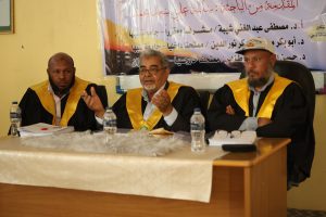 Three men in black robes with yellow sashes sit at a panel table during a conference, with an Arabic banner behind them.