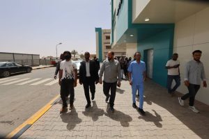 A diverse group of people walks along a paved sidewalk outside a modern teal and beige building, some in scrubs and others in casual or business attire.