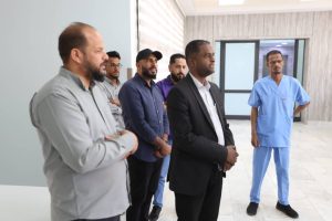 A group of men stands in a hospital-like hallway as a man in a black suit speaks, with a nurse in blue scrubs standing to the right.