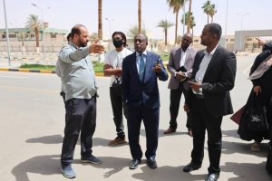 Man in a light gray shirt gestures while a group of professionals in suits and smart casual attire listen outdoors on a sunny street.