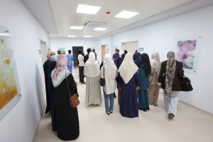 Group of people in traditional dress gathered in a bright hospital hallway with overhead lights and artwork on the walls.