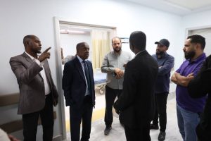 Group of men in business attire stand in a hospital corridor; one man points as they discuss near an open patient room.