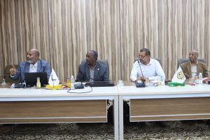 Four men in suits seated at a conference table with microphones, beverages, and papers in a formal meeting.]','