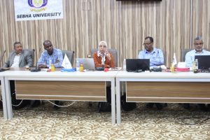 Five-person panel seated at a long conference table in a meeting room, with laptops, microphones, and drinks; a SEBHA University banner hangs on the wood-paneled wall behind them.