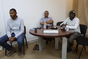 WhatsApp Image 2026-05-13 at 2.13.36 PM - جامعة سبها | Sebha University Three men sit around a round table in a meeting room, with cups, a water bottle, and papers on the table.