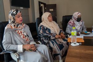 WhatsApp Image 2026-05-14 at 12.06.35 PM - جامعة سبها | Sebha University Three women in hijabs sit in office chairs around a conference table during a meeting, one wearing a face mask.