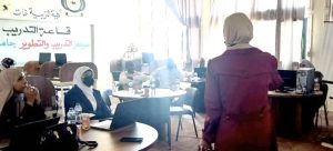 Instructor in a hijab addresses a group of women seated around round tables with laptops during a training session in a bright classroom.