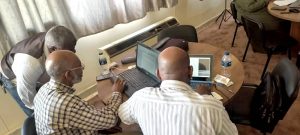 Three men sit around a round table, collaborating on open laptops during a meeting in an office.