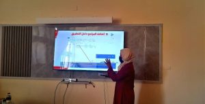 Woman in a hijab and maroon clothing giving a presentation beside a mounted screen displaying an Arabic slide in a classroom setting.