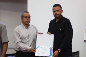Older man in a striped shirt hands a certificate to a younger man in a black shirt, both facing the camera at a ceremony edge.