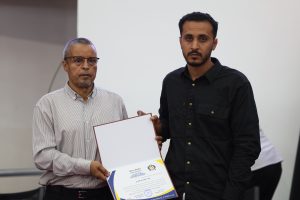 Two men stand side by side; one presents a certificate to the other at a formal event.