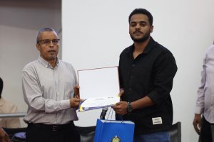Two men stand side by side as one presents a certificate to the other during a formal award event, with a blue gift bag visible in the foreground.