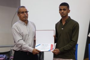 Two men stand side by side as an award certificate is handed over; the older man in a striped shirt and glasses presents it to a younger man in an olive shirt.