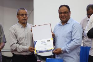Two men in striped shirts present a certificate at a formal event, with a blue gift bag visible nearby.