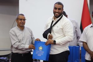 Two men exchange a blue gift bag at an indoor event, smiling for the camera.