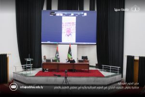 Auditorium stage with a large screen showing a video call of a man; two presenters sit at a desk with laptops, Libyan flags, and dark curtains behind.