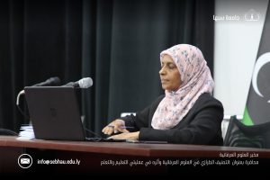 Woman wearing a pink floral hijab seated at a desk with a laptop and microphone during a conference talk.