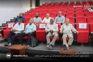 Group of men seated in a red-auditorium, watching a presentation in a lecture hall with Arabic logos visible on the right side.