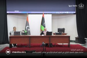Two presenters sit at a long wooden desk on a stage with Libyan flags and a large screen behind them, microphones in front of them (conference setting).