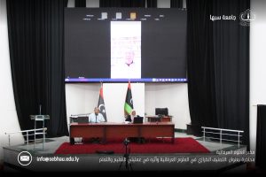 Lecture hall stage with a large screen showing a video call; two presenters sit at a desk with laptops, nearby Libyan flag and another flag.
