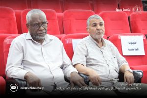 Two older men sit in bright red theater seats during a formal event, one in a white shirt and the other in a light plaid shirt, looking thoughtful.