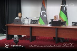 Two presenters sit at a long wooden table with laptops and microphones, Libyan flags behind them in a conference room, a camera on a stand foreground.