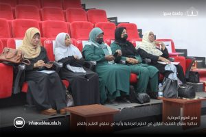 Women in hijabs seated in red theater seats during a conference, some looking at phones. A banner with Arabic text and a logo appears on the right side of the stage.