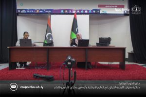 Panel discussion scene: a woman speaks into a microphone at a long desk with a man on a laptop beside her; Libyan flag and stage backdrop behind them.