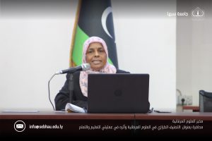 Female speaker wearing a pink hijab delivers a talk behind a podium with a laptop and microphone, with flags in the background and a watermark along the bottom edge.