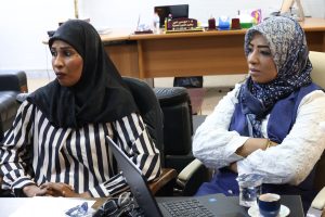 WhatsApp Image 2026-05-20 at 10.03.10 AM (1) - جامعة سبها | Sebha University Two women in hijabs sit at a conference table, one in a striped blouse and black scarf, the other in a blue patterned hijab, with laptop and papers nearby.