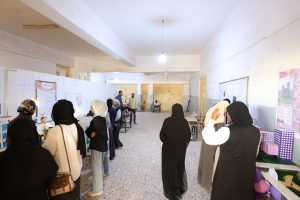 Indoor hall crowded with people in hijabs at display booths and posters on the walls, suggesting a community event or fair.