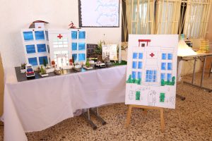 Colorful hospital-diagram diorama on a table with miniature figures, vehicles, and trees.