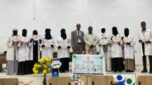 Group of students in white coats holding certificates on a stage, flanked by two men, with a banner and a flower arrangement in front.