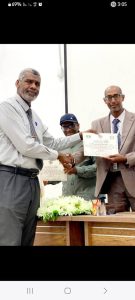Two men in formal attire shake hands as a certificate is exchanged at a recognition ceremony, with a flower-decorated table nearby.