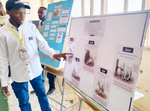 Young student in a white shirt points to a large poster about animals at a science fair.