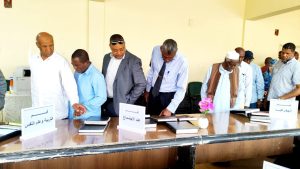 Group of men around a conference table reviewing documents; signs read Education and Psychology, Sociology, and Geography on the table centerpiece plant visible.