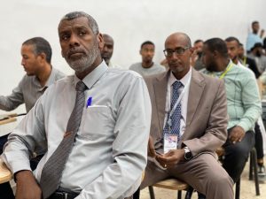 Man in a gray shirt and tie sits in the foreground of a classroom, with other attendees behind him in business attire.