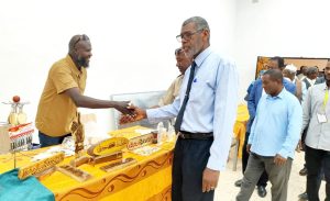 Man in a blue shirt and tie shakes hands with a vendor in a mustard shirt at a table of carved wooden items during an outdoor exhibition.