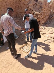 Group of young men in a rocky desert site examining artifacts laid out in a wooden tray on the ground. - They wear lanyards and casual clothes, walls of stones behind them.