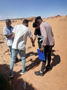 Four young men in a desert field lean over a wooden tray on sand, examining small objects with ID lanyards on their necks.