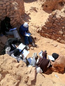Group of women in hijabs sitting in a sandy ruin, taking notes on clipboards.