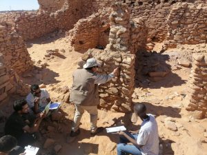 Archaeologists study a desert ruin, some taking notes on clipboards near a stacked-stone wall encampment.