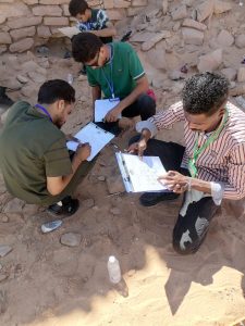 Four men in casual clothes with clipboards and lanyards conduct a field survey in a rocky desert area, kneeling and writing notes near a stone wall.