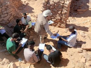 Group of students in a rocky desert site, seated in a circle, taking notes while an instructor with a wide hat guides them.