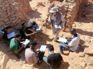 Group of researchers sitting among rocky desert ruins, taking notes as a guide explains with a clipboard.
