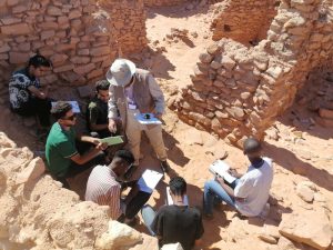Group of researchers crouched in a sandy ruin, taking notes on clipboards under bright sun.