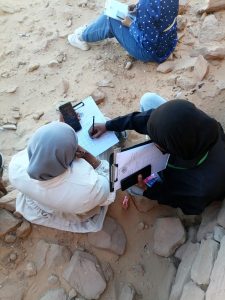 Three people in hijabs sit on rocky desert ground, writing on clipboards and checking a smartphone during field data collection.