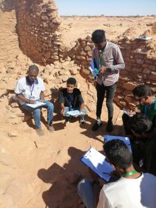 Group of students at a desert excavation site, notebooking and sketching among stone walls while wearing name badges.